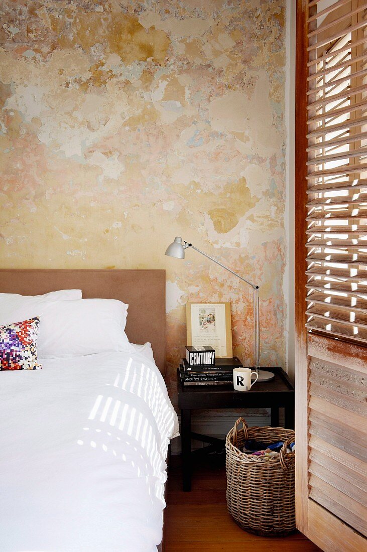 Bed with white bed linen, bedside table and laundry basket in front of wall with vintage patina