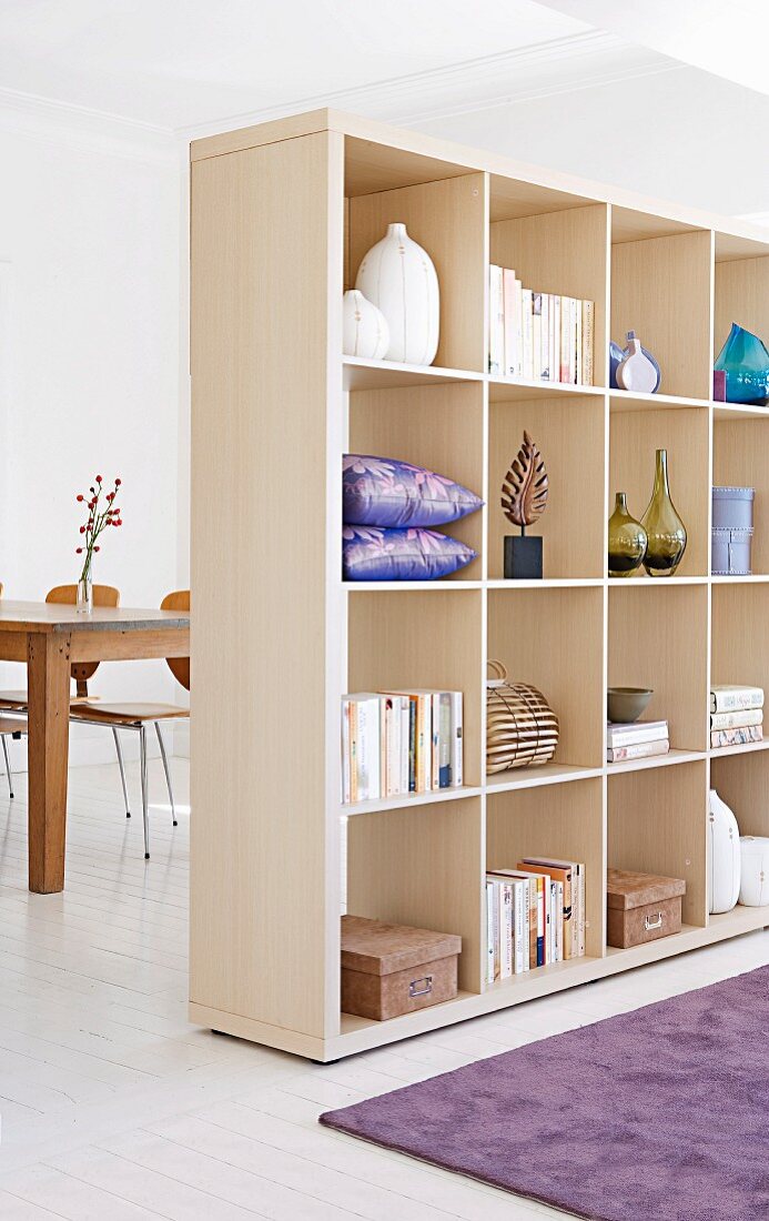 Pale, wooden partition shelving filled with books, vases and cushions; dining area in background