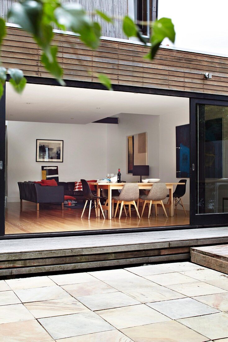 Blick von plattenbelegter Terrasse durch weit geöffnete Glasschiebetüren in offenen Wohnraum