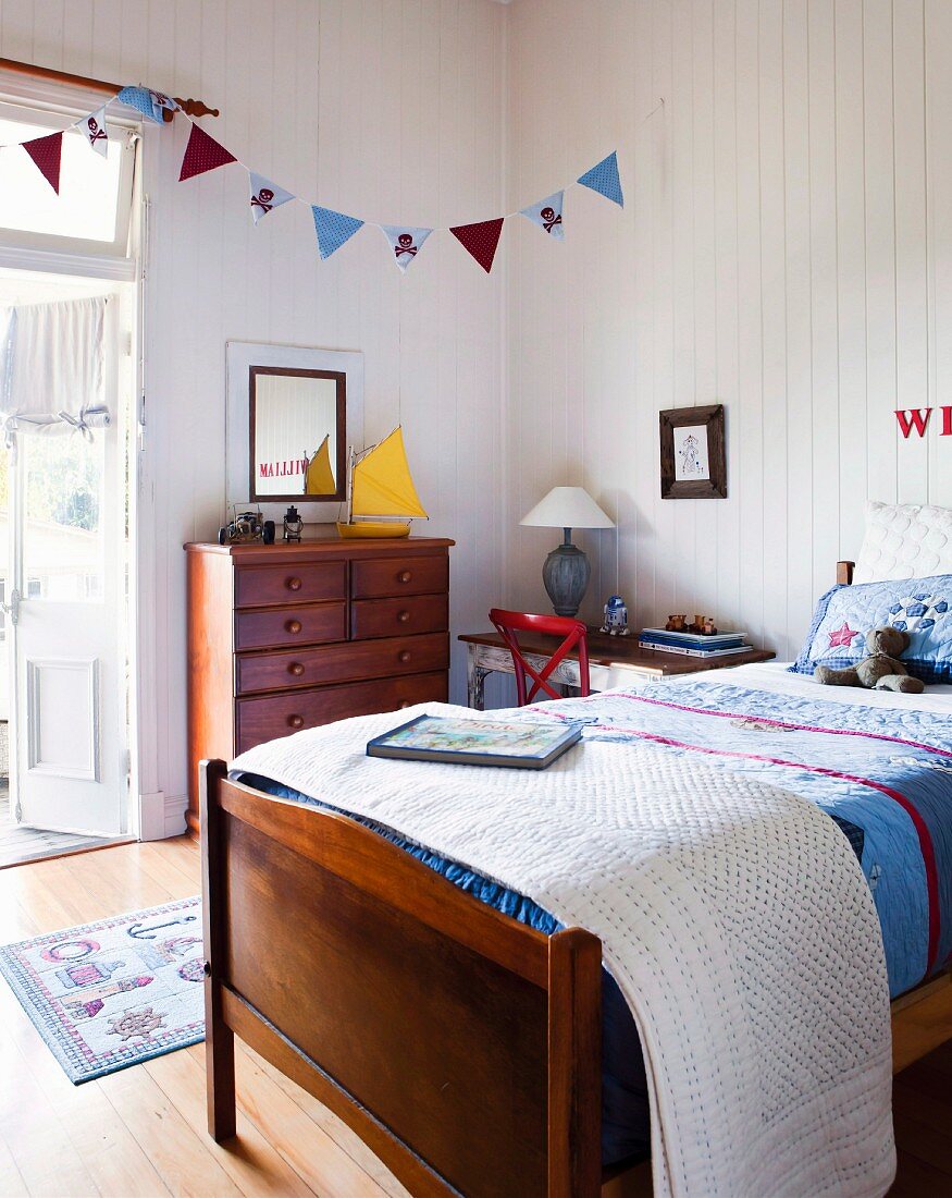 Bett mit Patchworkdecke, Wimpelgirlande über Holzkommode und weiße Holzvertäfelung in freundlichem Landhaus-Kinderzimmer