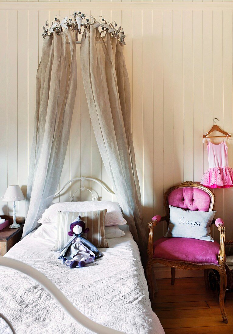 Stoffpuppe auf romantischem Kinderbett mit Kronen-Baldachin, daneben ein pinkfarben bezogener Antikstuhl