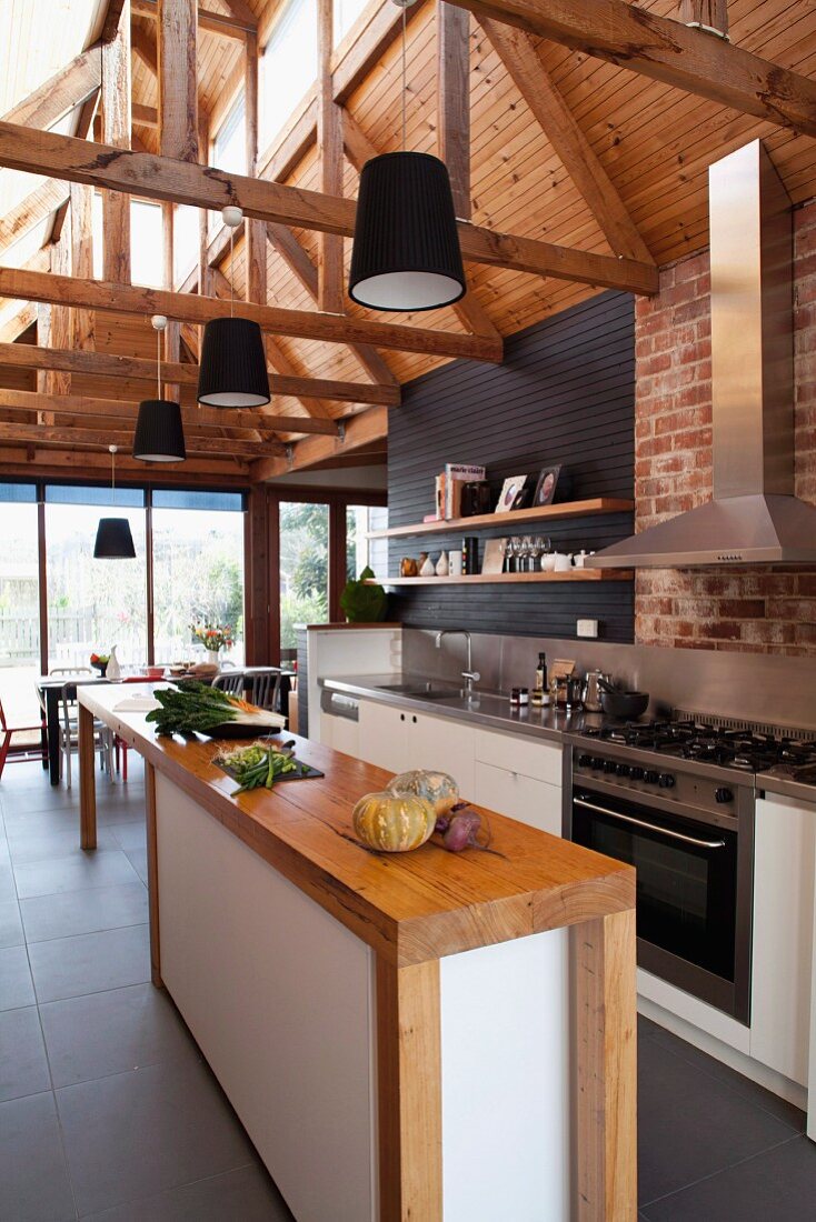 Schmale Holztheke mit Schrankunterbau in offener hoher Designerküche; sichtbare Dachkonstruktion mit montierten schwarzen Pendelleuchten