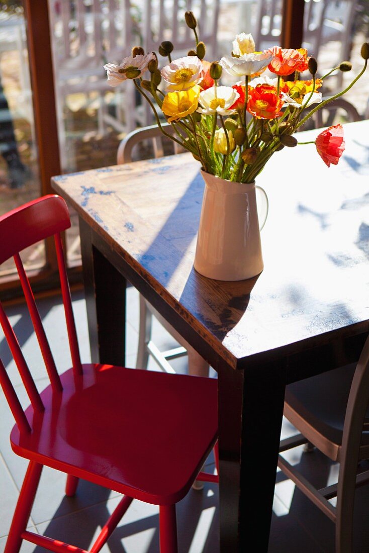 Verschiedenfarbiger Mohnblumenstrauss in Porzellankrug auf sonnigem Vintage-Tisch, davor rot lackierter Küchenstuhl