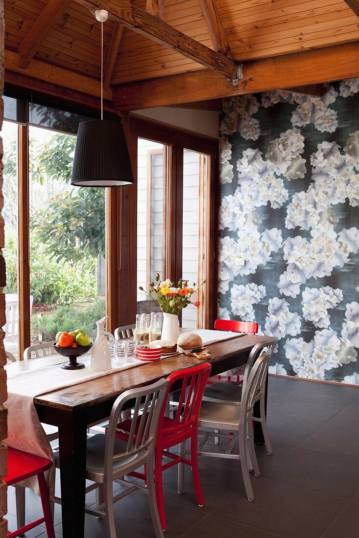 Red and white chairs at rustic wooden table in front of wall covered in floral wallpaper