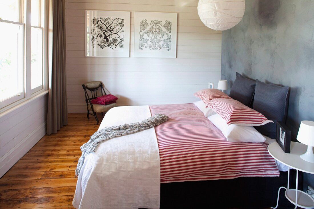 Doppelbett mit rot-weiss gestreifter Bettwäsche an grauer Wand, im Hintergrund weiße Holzwand in ländlich modernem Schlafzimmer
