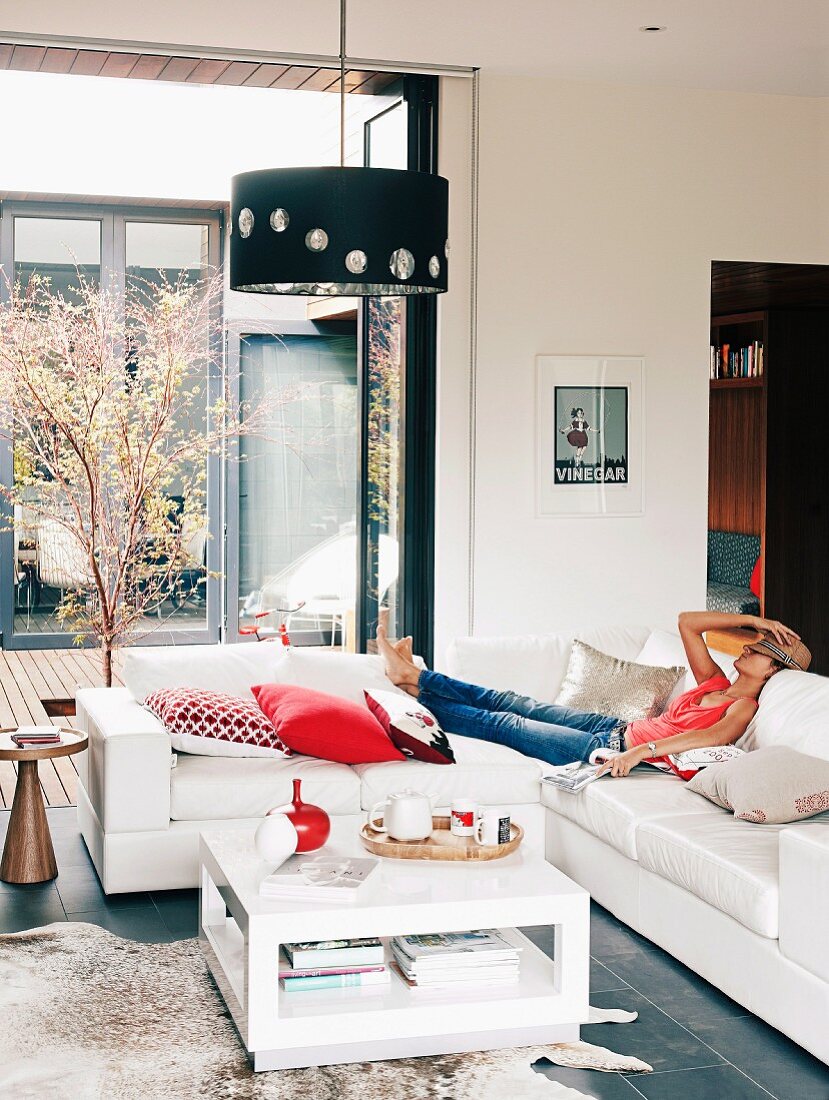 Bright living room with folding sliding doors to courtyard; young woman relaxing on corner sofa with scatter cushions
