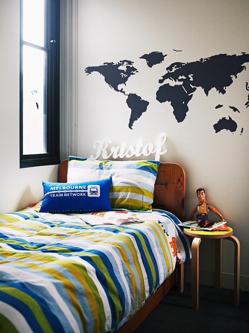 Teenager's bed with striped covers and Alva Aalto wooden stool below world-map stickers and name on wall