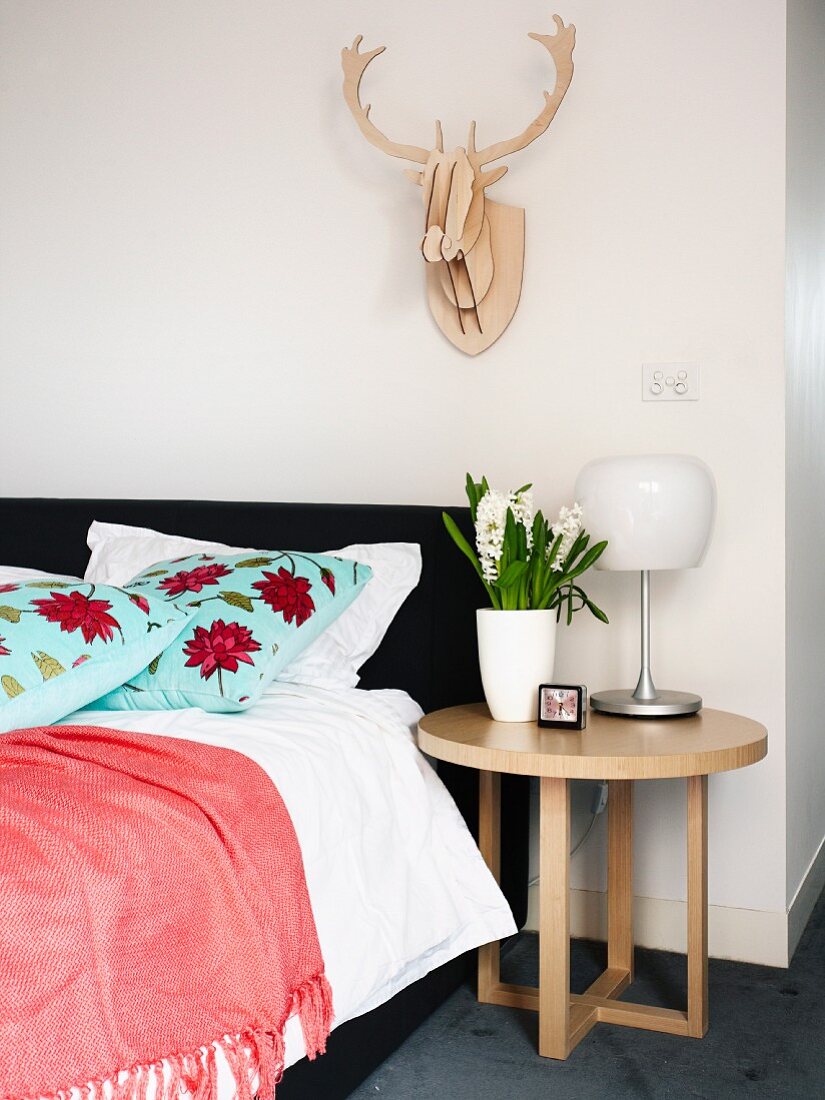 Runder Beistelltisch aus Eichenholz neben Polsterbett mit dekorativ geblümten Kissen und stilisiertes Hirschgeweih an der Wand