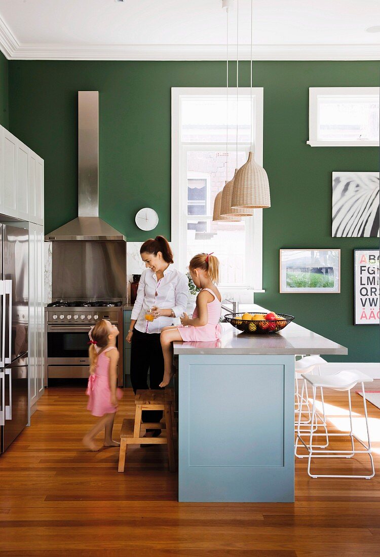 Mother and children in open-plan kitchen with little girl sitting on counter and stainless steel cooker against dark green wall in background