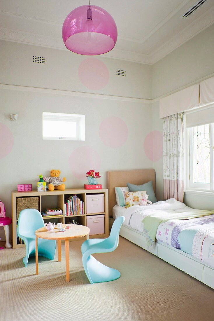 Kinderzimmer mit hellblauen Schalenstühlen aus Kunststoff um runden Holztisch, in Zimmerecke Bett an Wand mit pastellfarbenen Kreisen