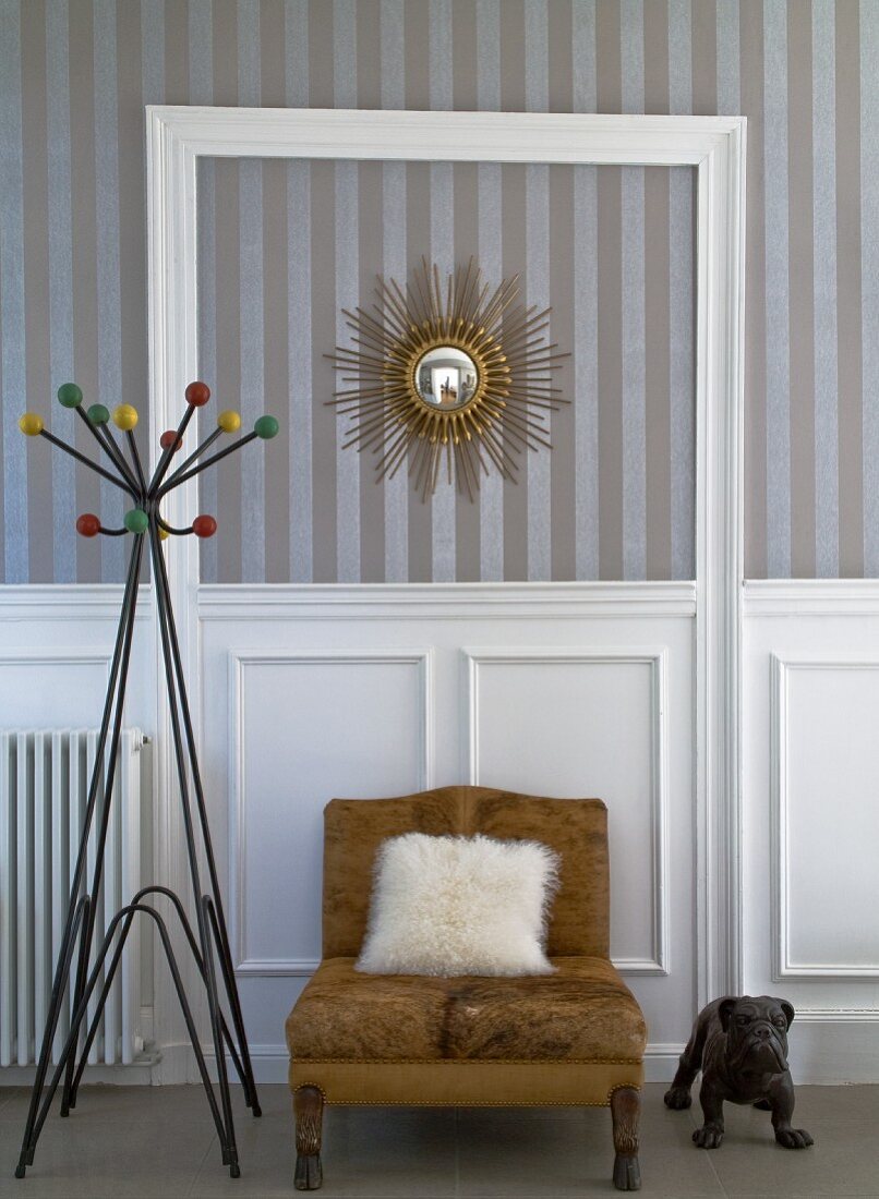 Fellkissen auf braunem Sessel und Retro Garderobenständer vor Wand mit weisser, halbhoher Holzverkleidung, darüber Streifentapete