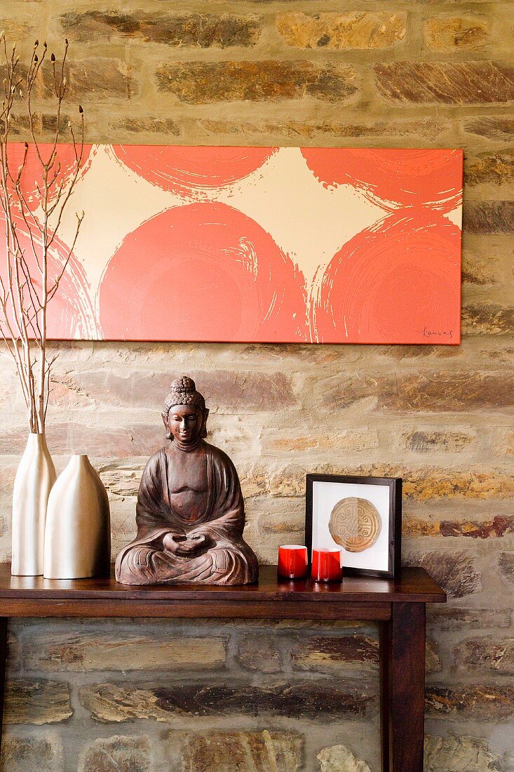 Buddhafigur zwischen Metall Vasen und Teelichtern auf dunklem Holztisch vor Natursteinwand mit modernem Bild
