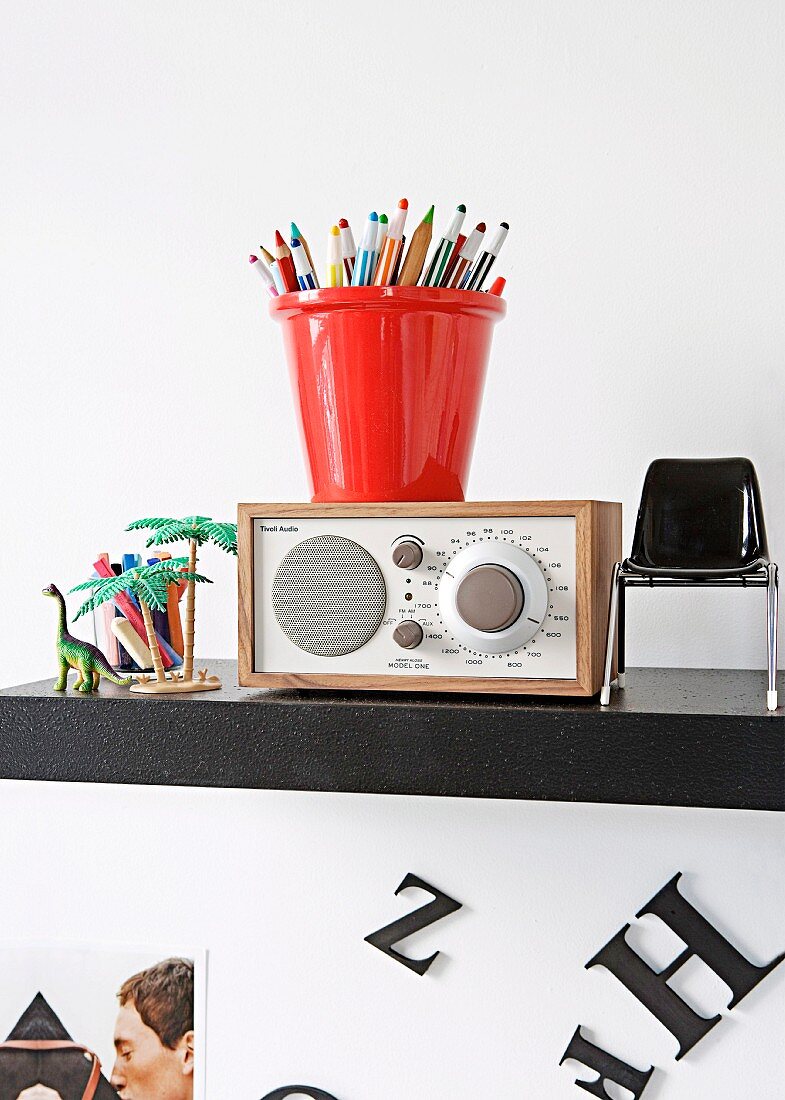 Buntstifte in rotem Behälter auf Radio zwischen Miniatur Stuhl und Spielzeug auf schwarzem Regal an Wand
