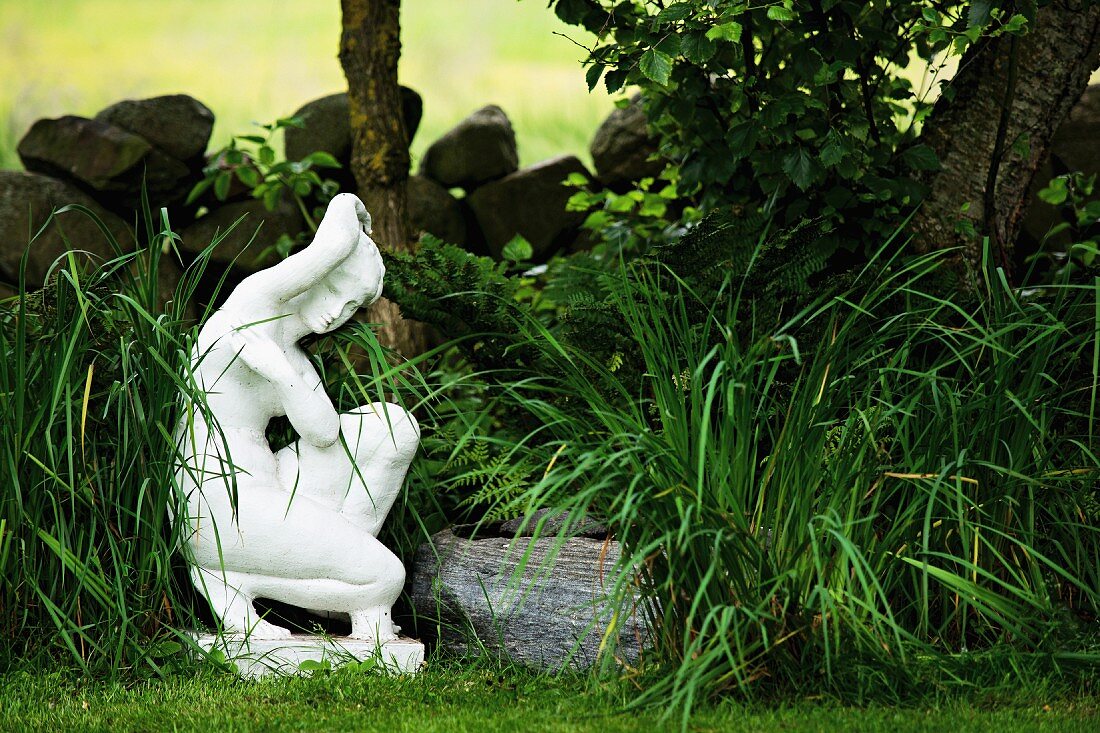 weiße Frauenfigur zwischen Gräsern und Steinmäuerchen im sommerlichen Garten