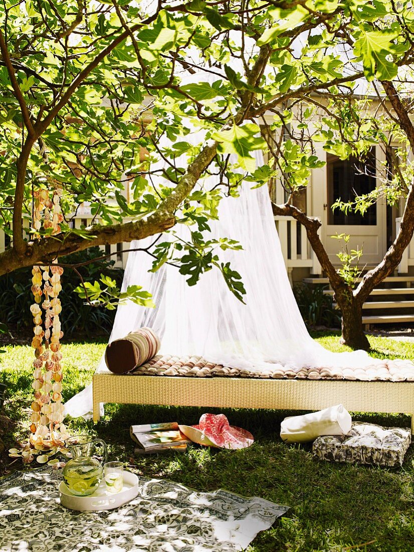 Schattiges Plätzchen im Garten - luftiger Baldachin über Tagesbett aus Rattan, davor Picknickdecke und Kissen in der Wiese