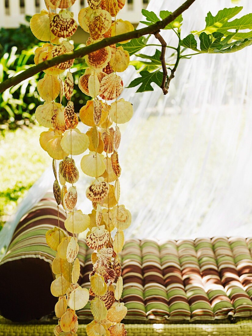 Ruheoase in sommerlichem Garten - aufgefädelte Muscheln an Baum aufgehängt, dahinter Liegepolster mit Streifenbezug in der Wiese