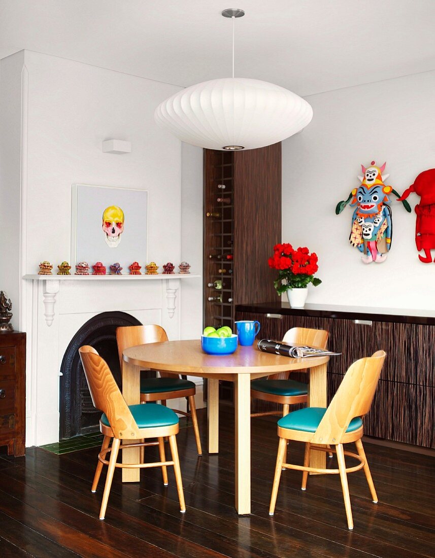 Pale, wooden dining set with turquoise seat cushions in front of dark wine rack and sideboard; colourful artworks on walls