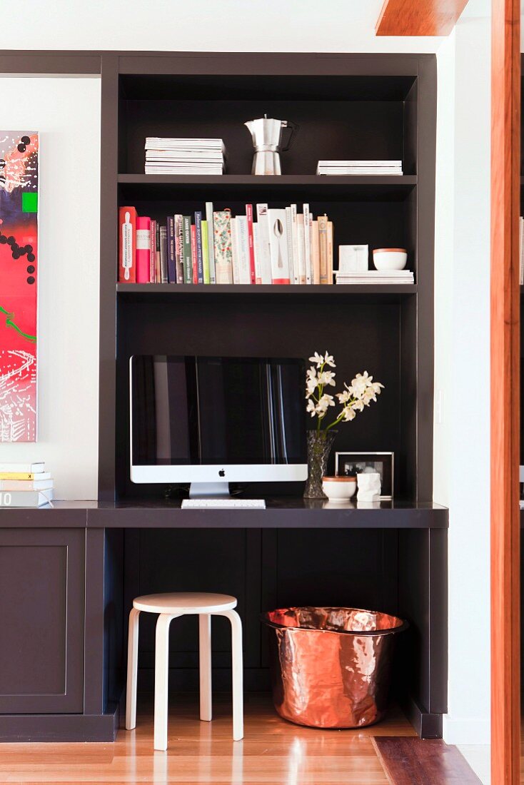 Small home office in dark fitted shelving niche with Scandinavian wooden stool and hammered copper pot as waste paper basket