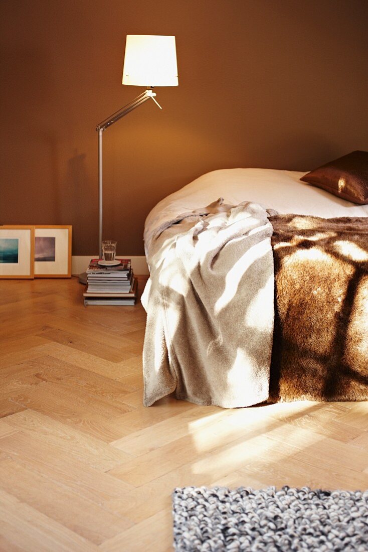 Modern, designer standard lamp and stack of books next to bed with fur blanket against brown-painted wall