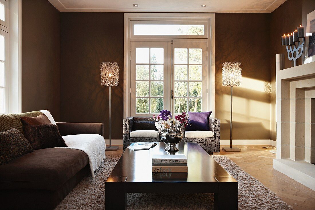 Glasflügeltür, flankiert von Stehlampen in braun getöntem Wohnraum mit eleganter Sitzgruppe vor dem Kamin