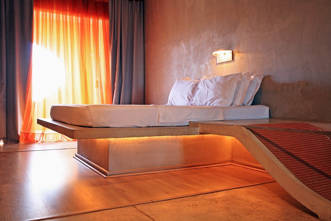 Futuristisches Schlafzimmer mit hinterleuchtetem Bett und angeschlossener Badewanne aus Beton, im Hintergrund transparenter Vorhang am Fenster