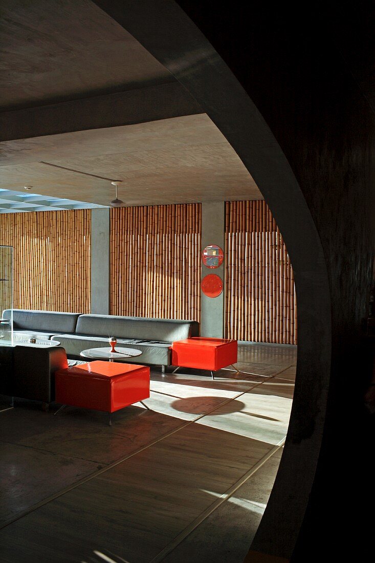 View though rounded opening into minimalist lounge area with grey and orange sofa set; screens of bamboo poles in windows in background