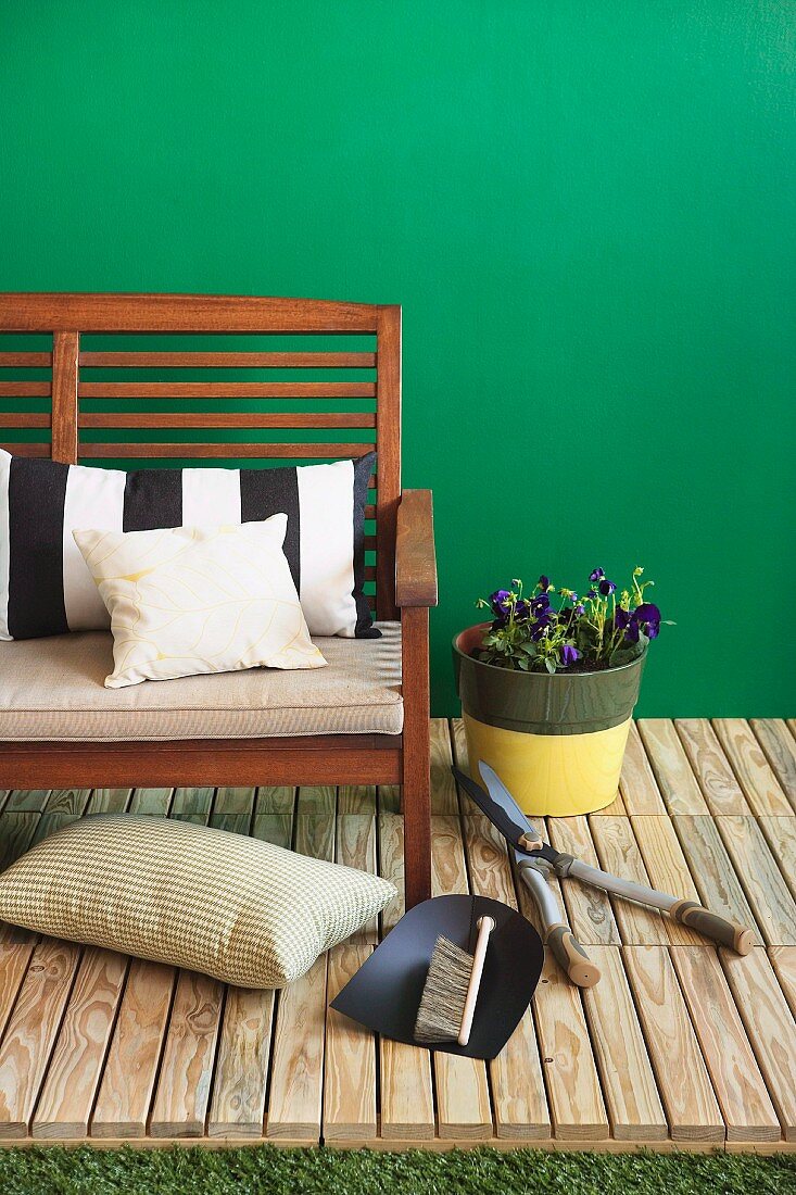 Potted plant next to garden bench and various gardening tools arranged on wooden decking against green-painted wall