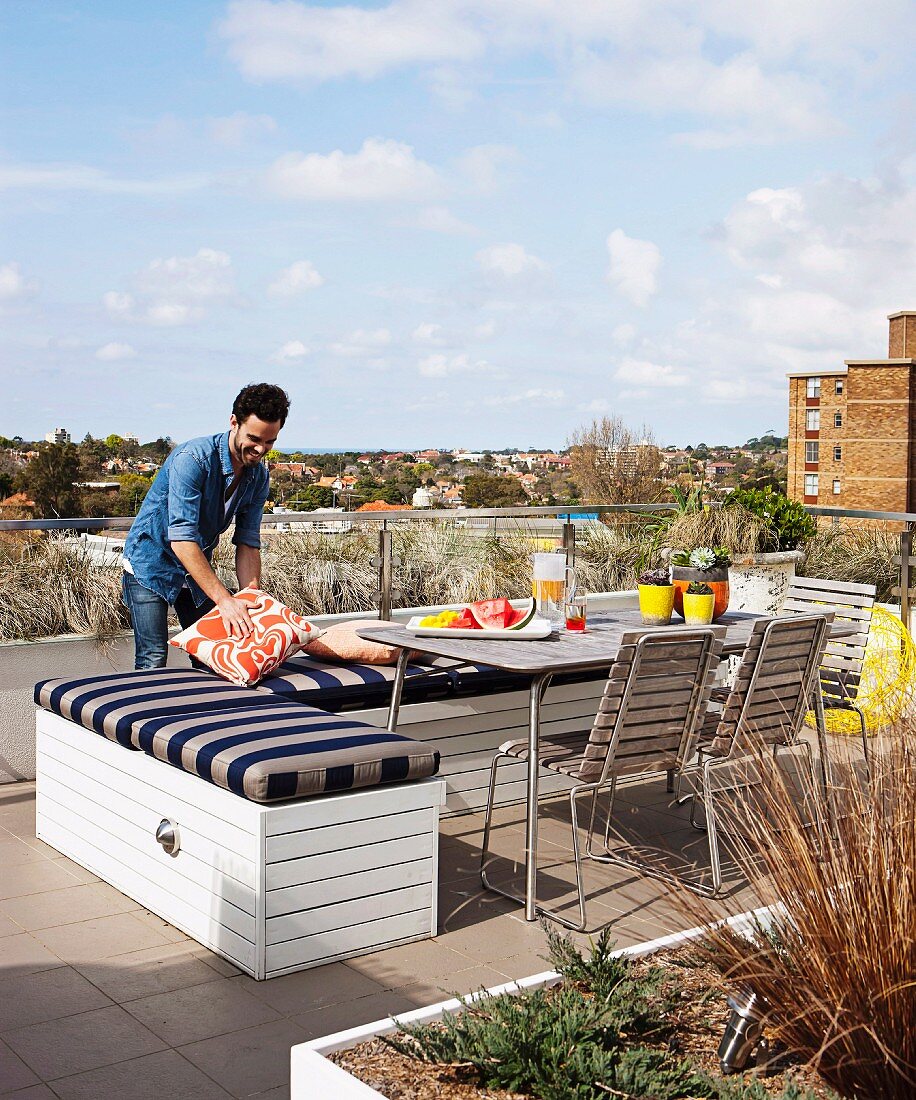 Bench with striped seat cushion, table and chairs on roof terrace; man arranging cushions