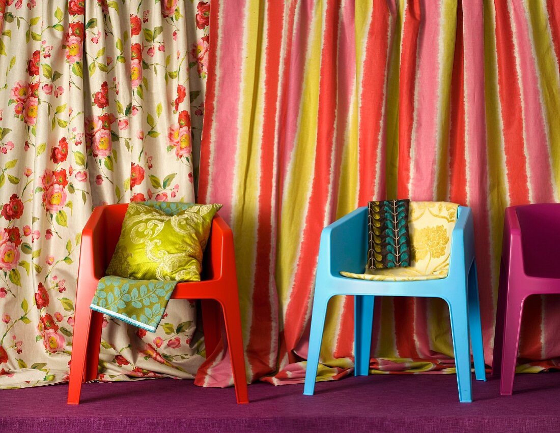 Scatter cushions and fabrics on colourful shell chairs; draped bolts of striped and floral fabrics in background