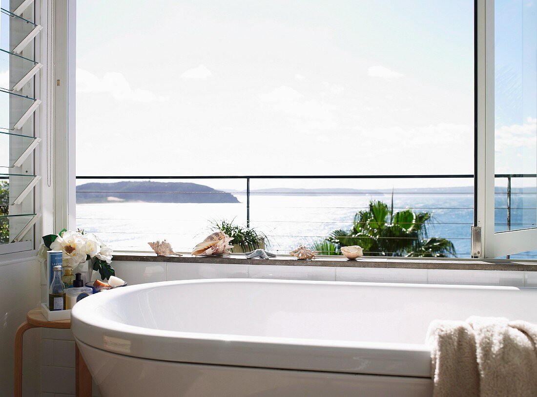 Bathroom with glorious sea view though open, folding glass window; seashell ornaments on edge of bathtub