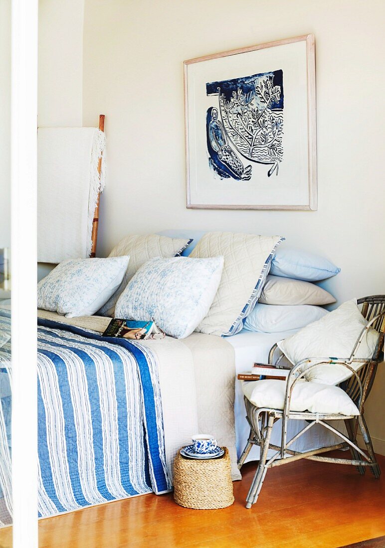 Korbstuhl vor französischem Bett mit Kissenstapeln und blaugestreifter Tagesdecke, darüber ein gerahmtes Bild