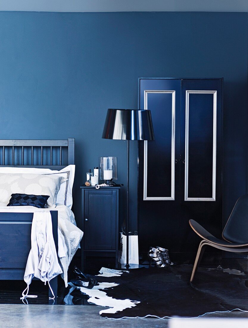 Blaue Stunde - Stehleuchte mit glänzendem Schirm zwischen Bett und Schrank; teilweise sichtbarer Klassikersessel auf Kuhhaut