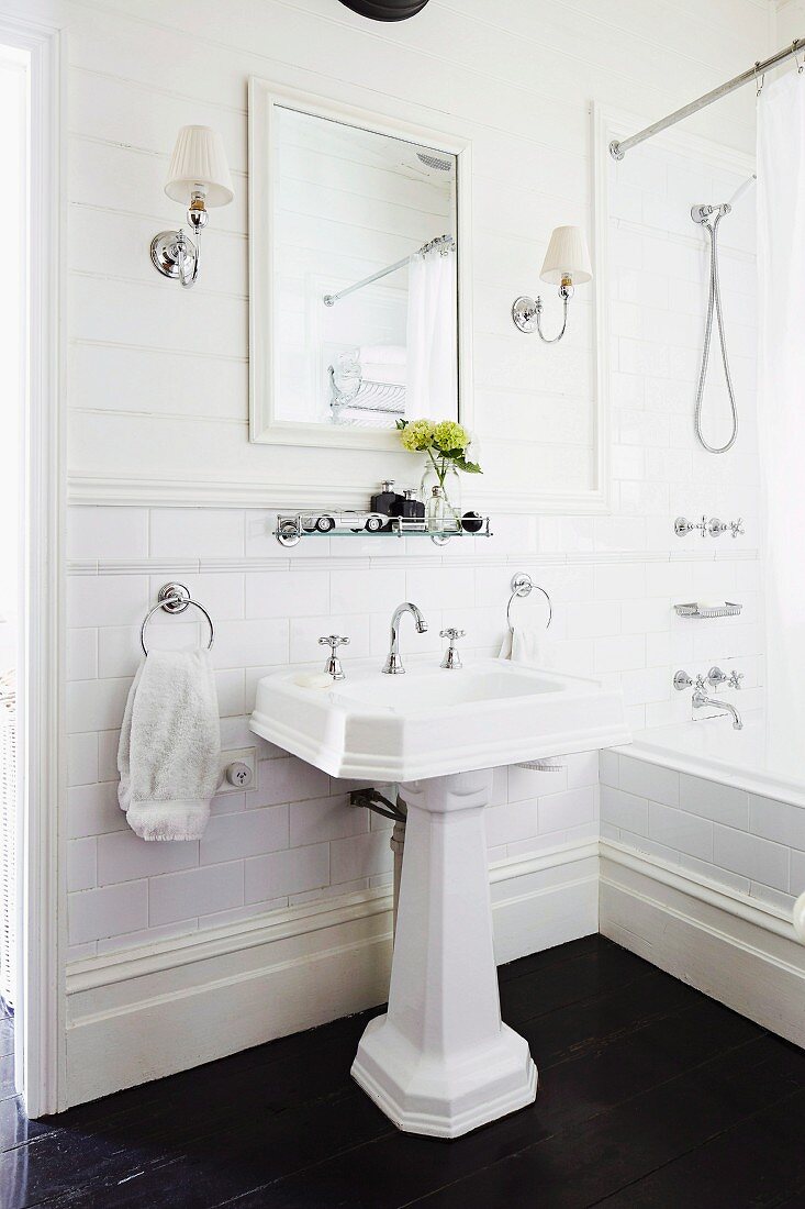 Vintage Standwaschbecken auf dunklem Dielenboden, gerahmter Spiegel, symmetrische Leuchten an weisser Holzwand in traditionellem Bad