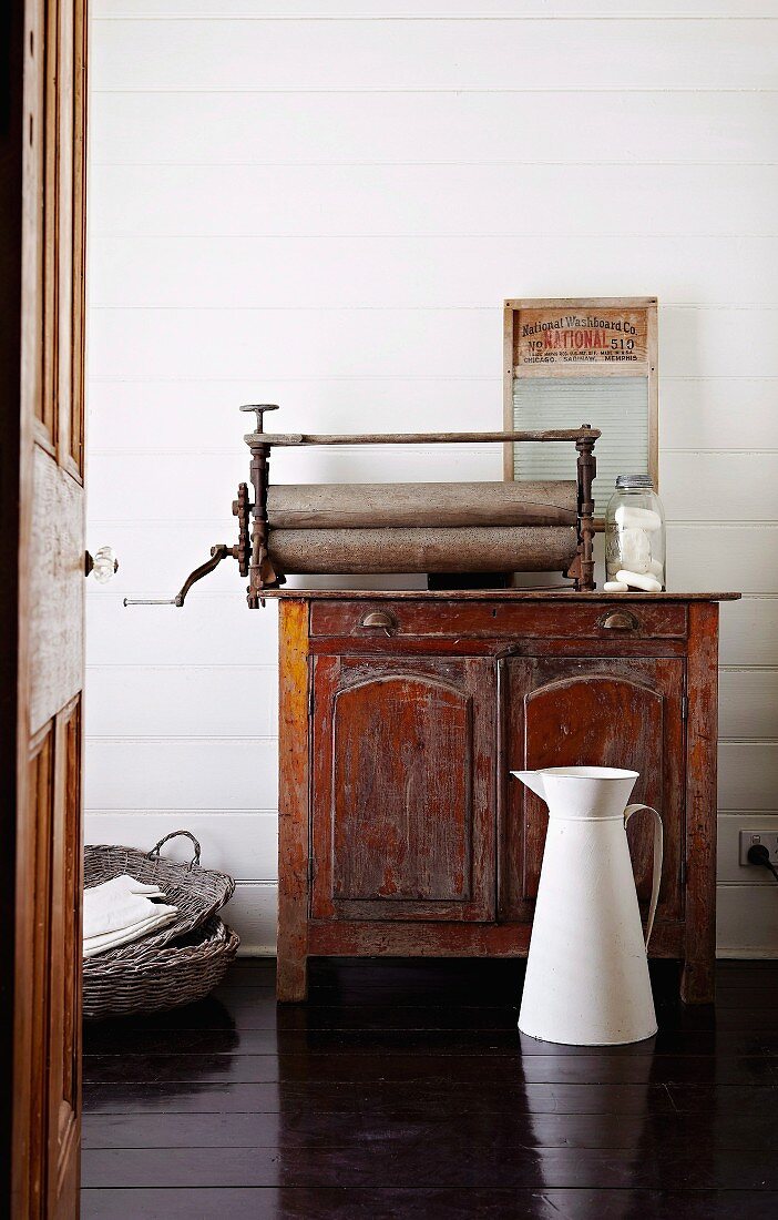 Blick auf weiße Metall-Wasserkanne auf dunklem Dielenboden und antiker Holzanrichte mit Vintage-Wäschemangel