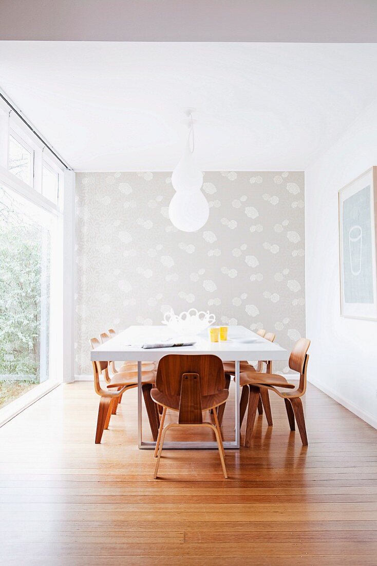 Weisser Esstisch mit Klassiker Holzstühlen in minimalistischem Zimmer, Stirnwand mit gemusterter Tapete, seitlich Glasfassade