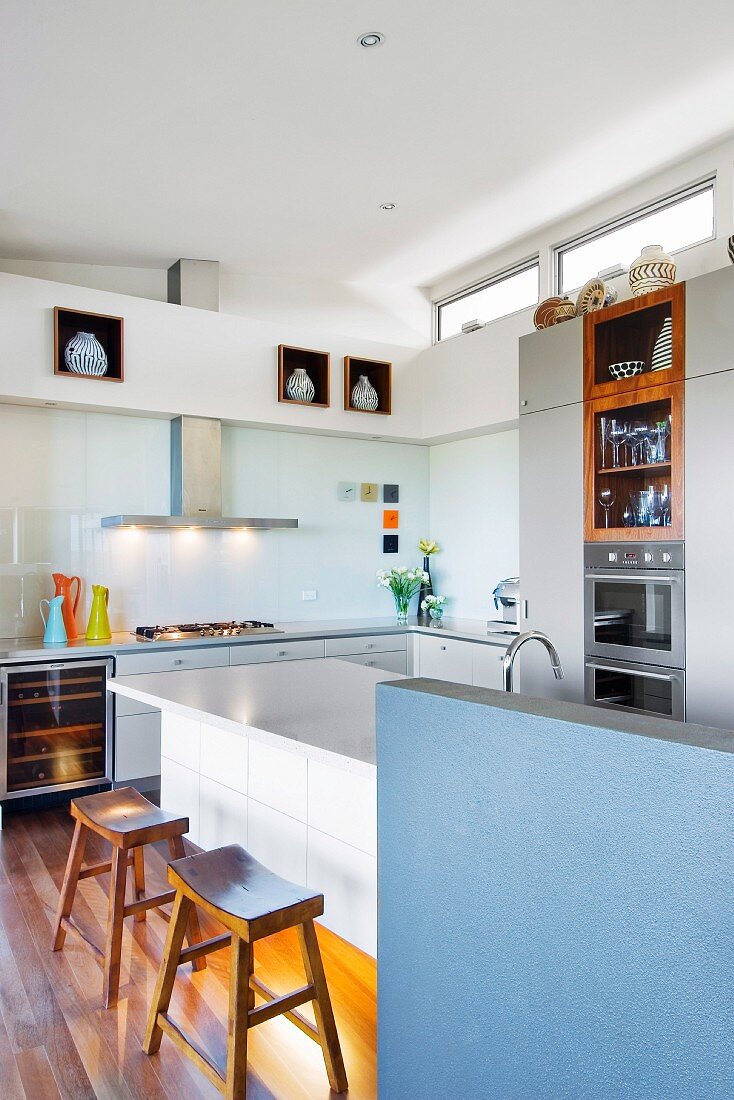 Kitchen island with blue, half-height wall and wooden stools in corner of open-plan kitchen with transom windows