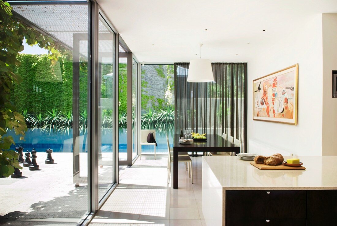 Offene Küche und Essplatz vor umlaufenden Glasfronten; Blick auf sonnige Terrasse und Pool mit begrünter Rückwand
