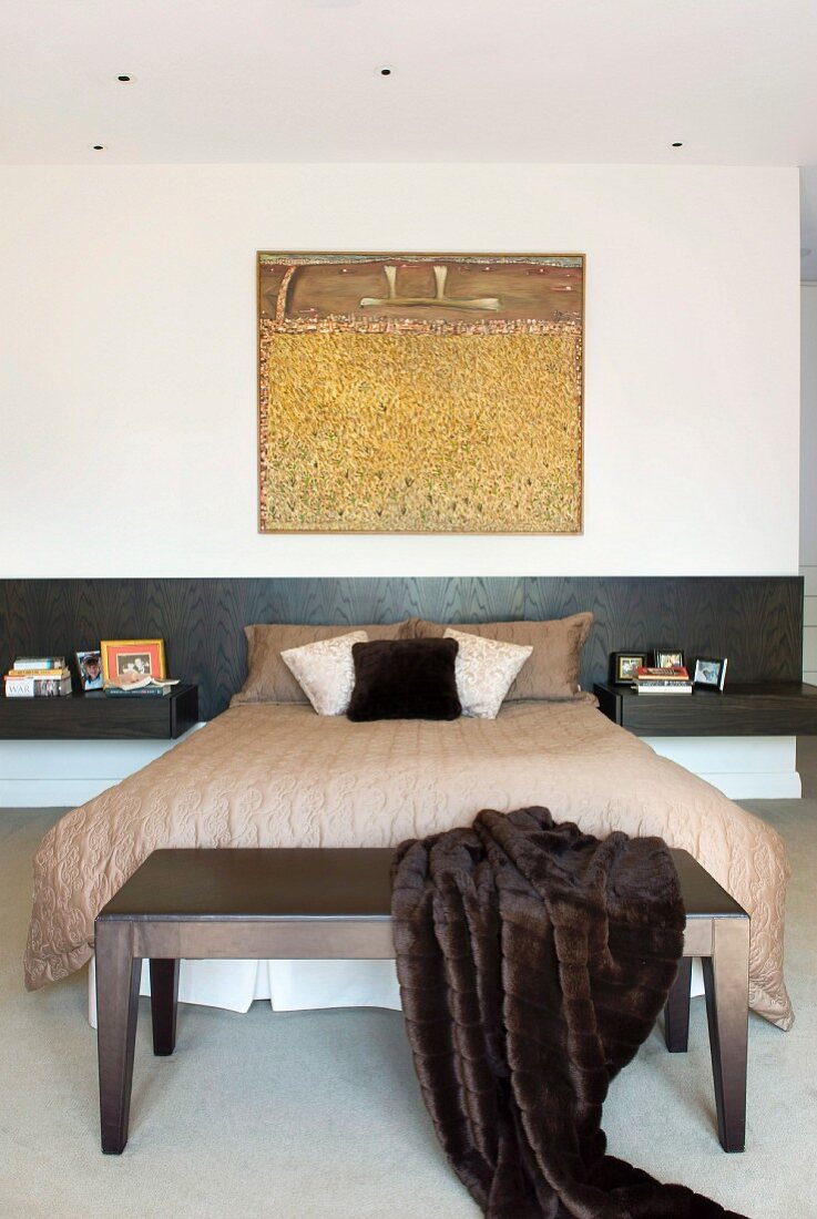 Modern artwork above French bed with black headboard and integrated bedside tables; fur blanket on bedroom bench in foreground