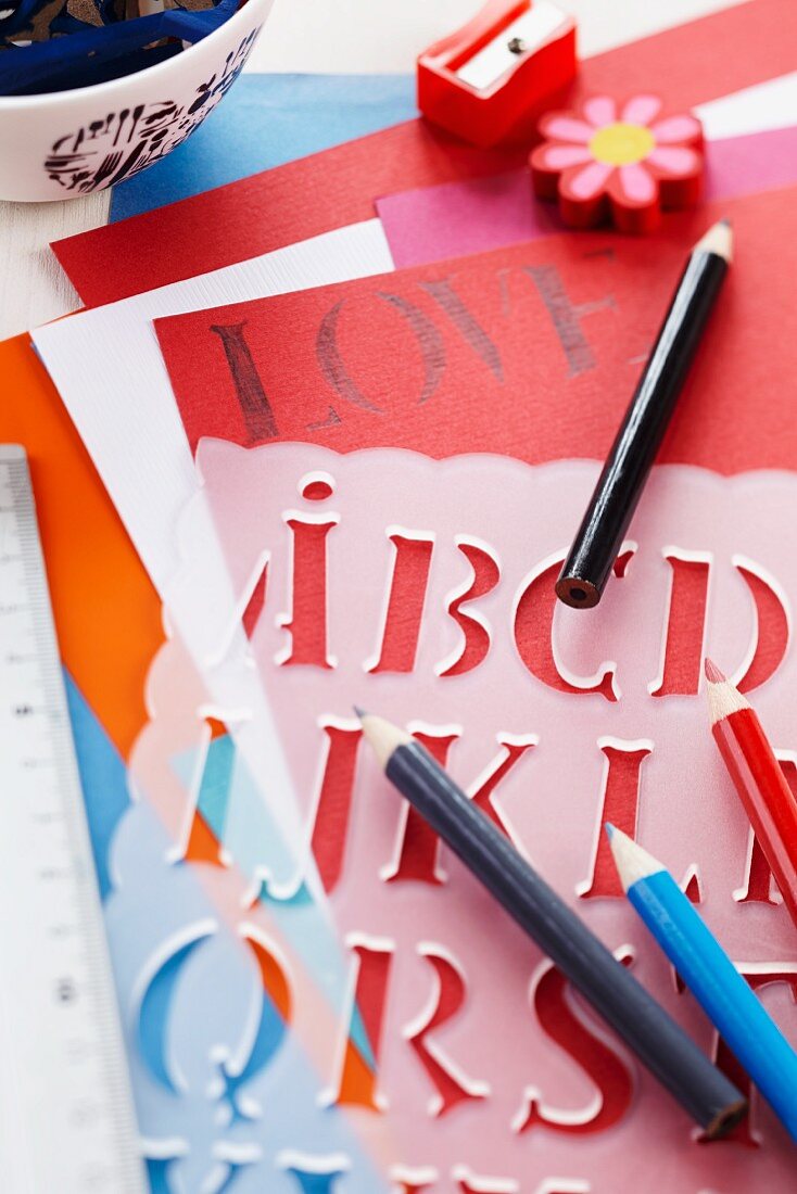 Buchstabenschablone aus Kunststoff und Stifte auf bunten Papierbögen