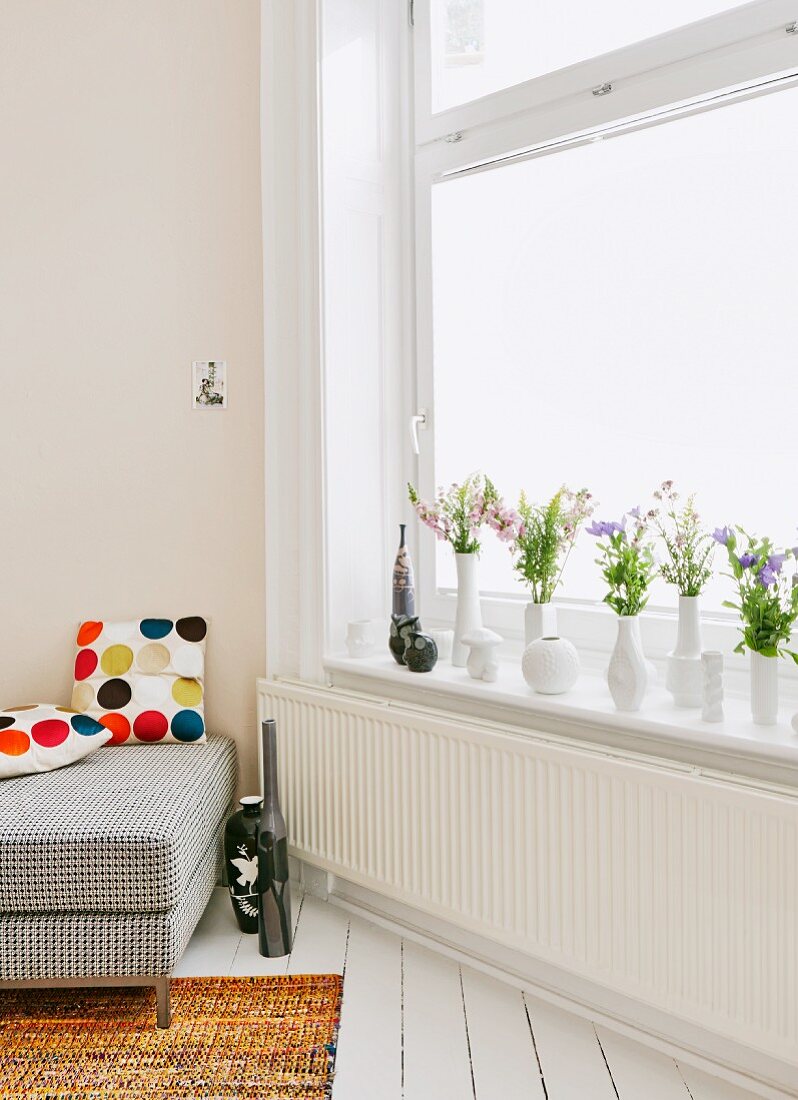 Flowers in collection of white vases on windowsill; partially visible sofa with scatter cushions to one side
