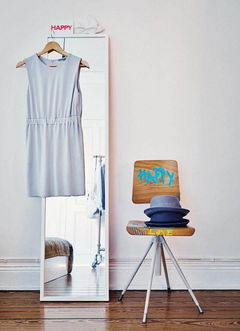 Stack of hats on painted, Danish, architect-designed chair next to dress on coat hanger hung on mirror