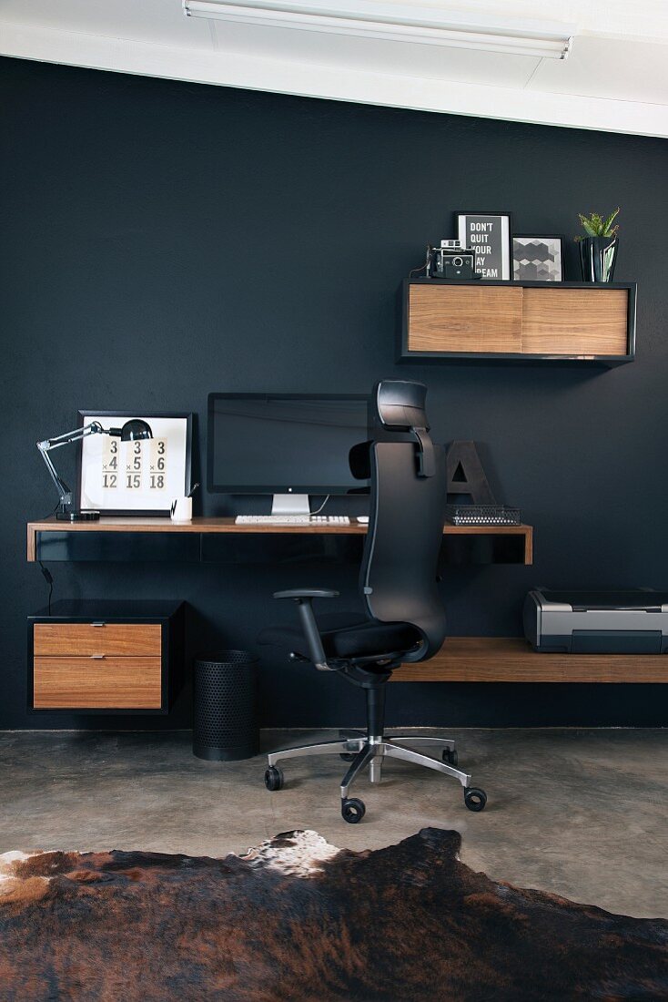 Homeoffice mit schwarzem Bürostuhl vor Arbeitskonsole zwischen kleinen Schrankmodulen an schwarzer Wand befestigt
