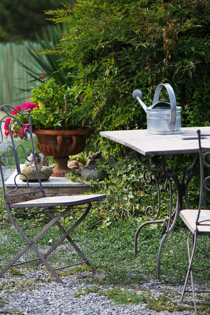 Gartentisch mit Giesskanne und Gartenstühle vor Strauch und Blumenamphore