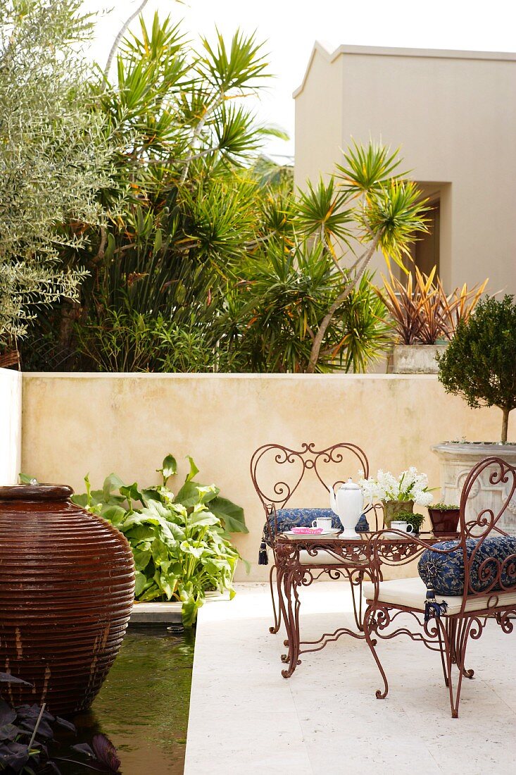 Delicate, curved metal chairs on stone terrace in Mediterranean surroundings with urn-shaped fountain in pool