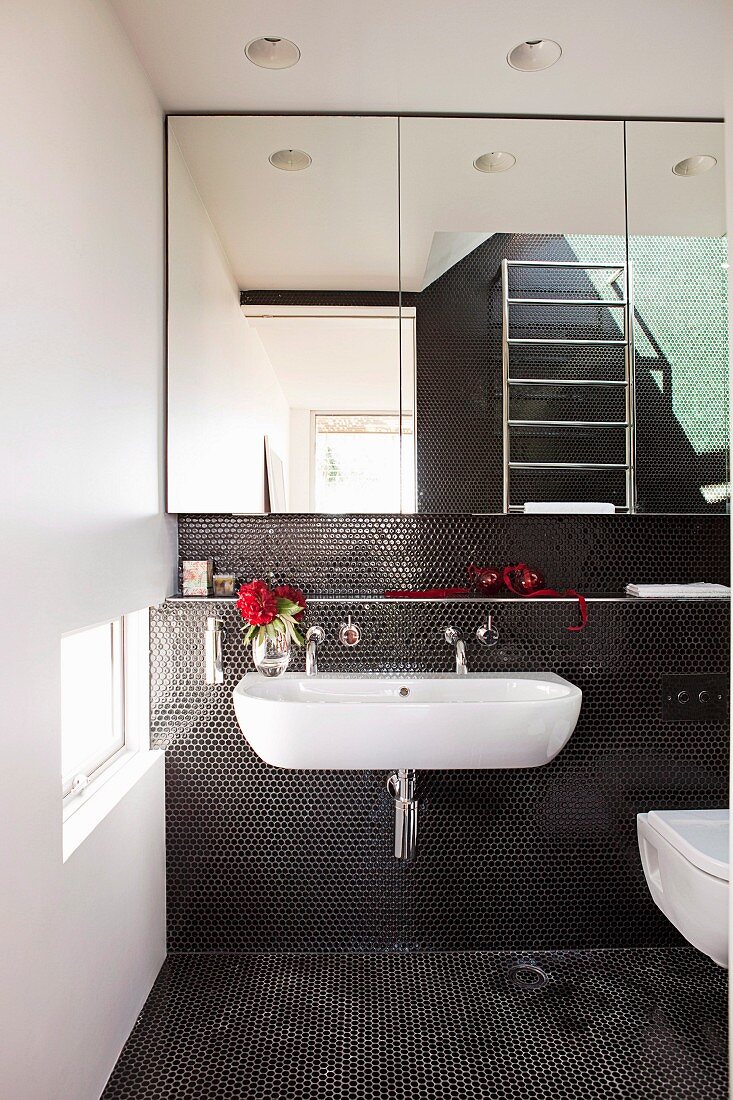 White sink on wall with black mosaic tiles below mirrored cabinets