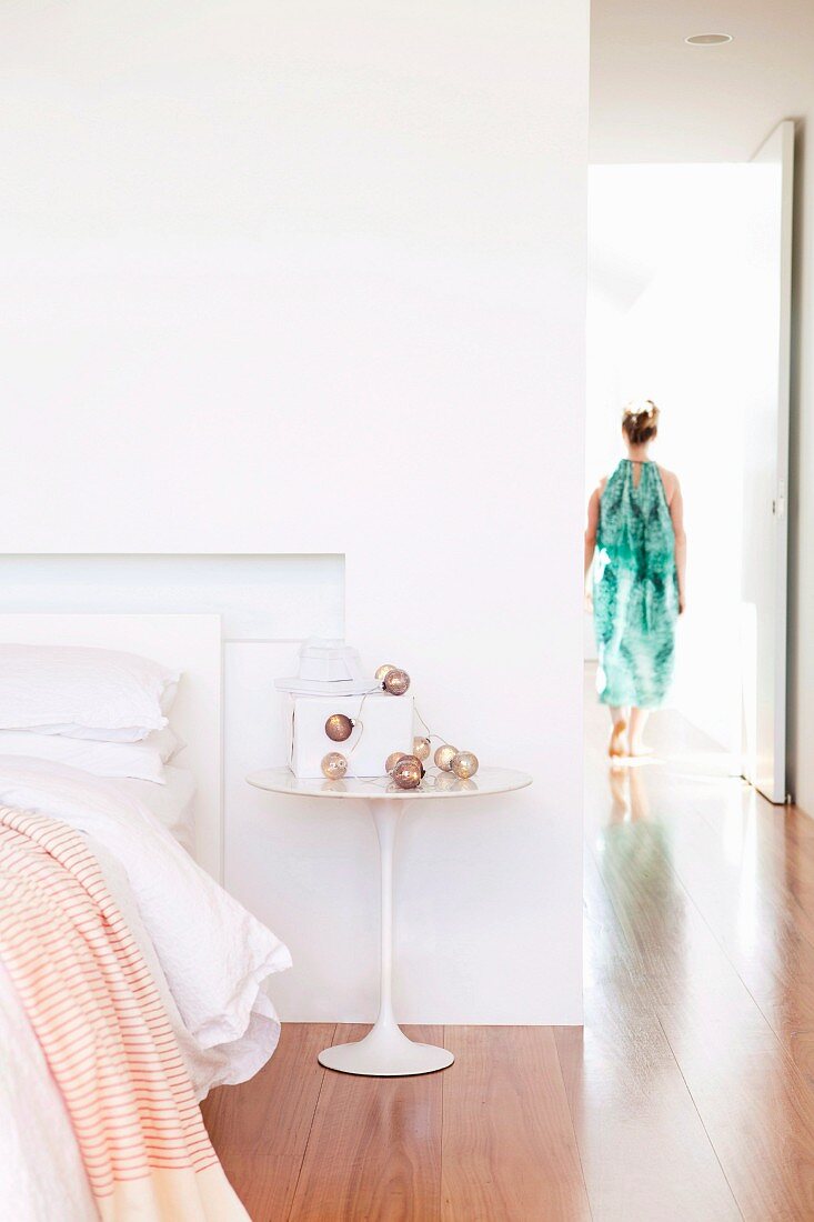 White, classic side table next to bed in open-plan bedroom; woman in hallway in background