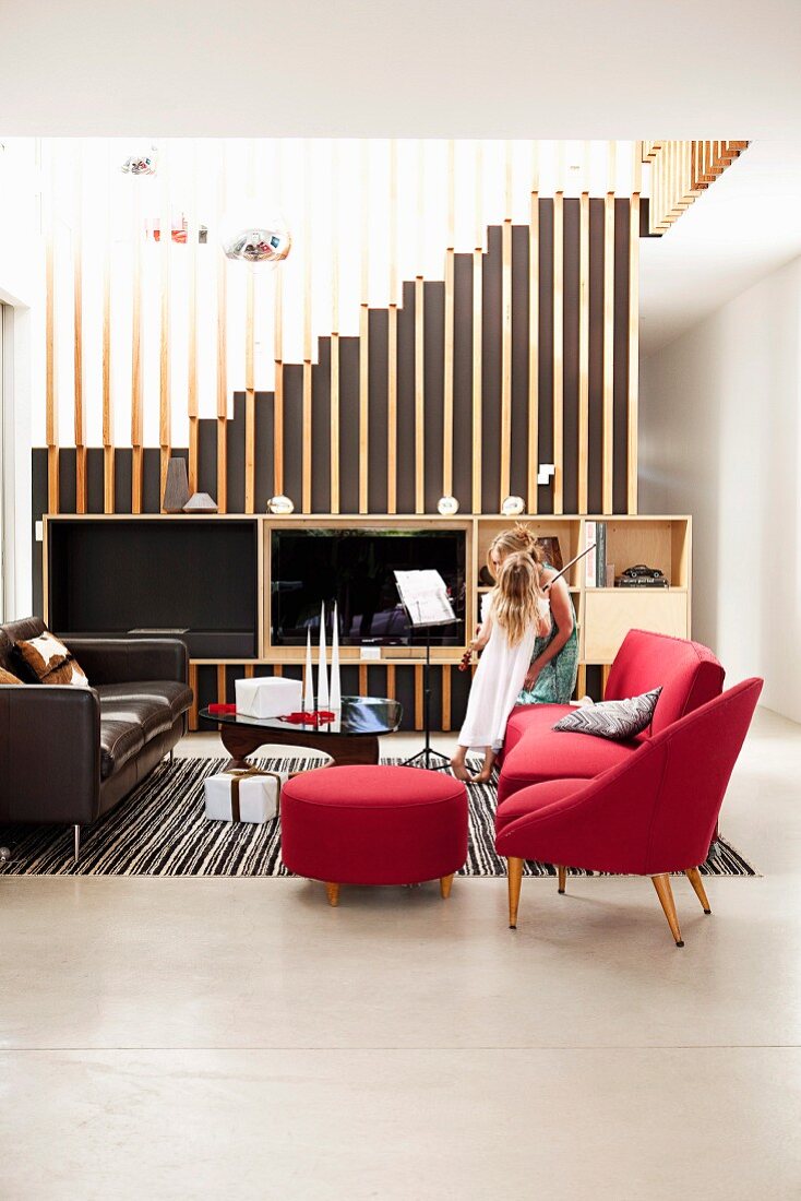 Red armchair with footstool and sofa in open-plan living area; media cabinet against staircase with floor-to-ceiling wooden slats as see-through partition