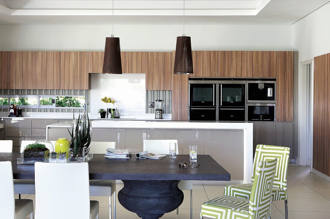 Offene Designerküche mit ruhiger Farbgestaltung in Grau, Weiß und holzfarbenen Elementen im Oberschrankbereich; Esstisch mit Säulenfuss und Polsterstühle im Vordergrund