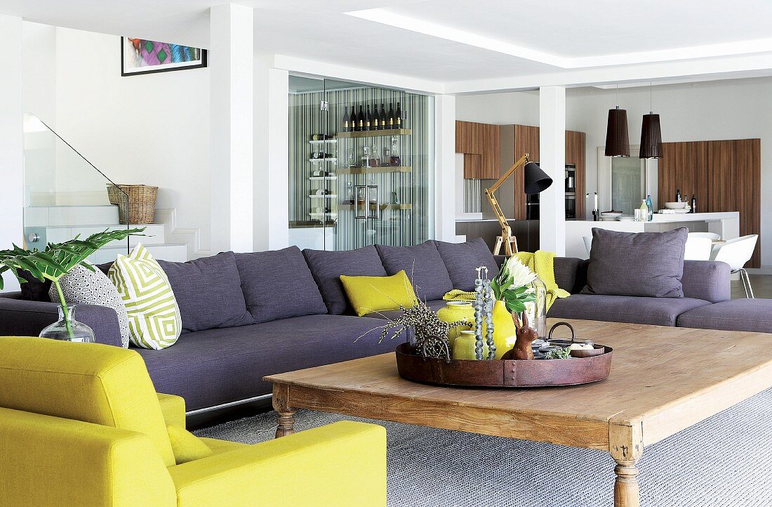Sofalandschaft in Grau und gelbgrün mit grossem Holztisch in offenem Wohnraum mit Treppe, Bar und Küche im Hintergrund