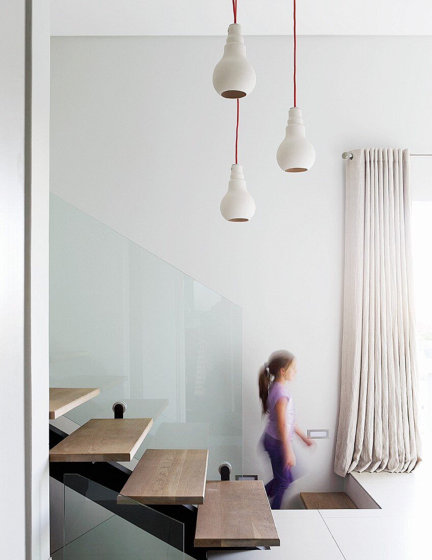 Pendant lamps above modern interior staircase made from steel girders with floating wooden treads and glass balustrade; girl in background
